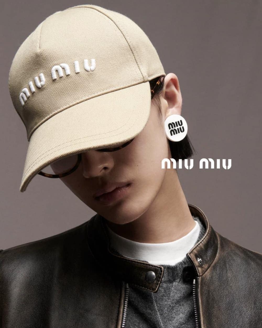 Fashion portrait of a person wearing a khaki Miu Miu cap, earrings, eyeglasses and leather jacket against a grey background.