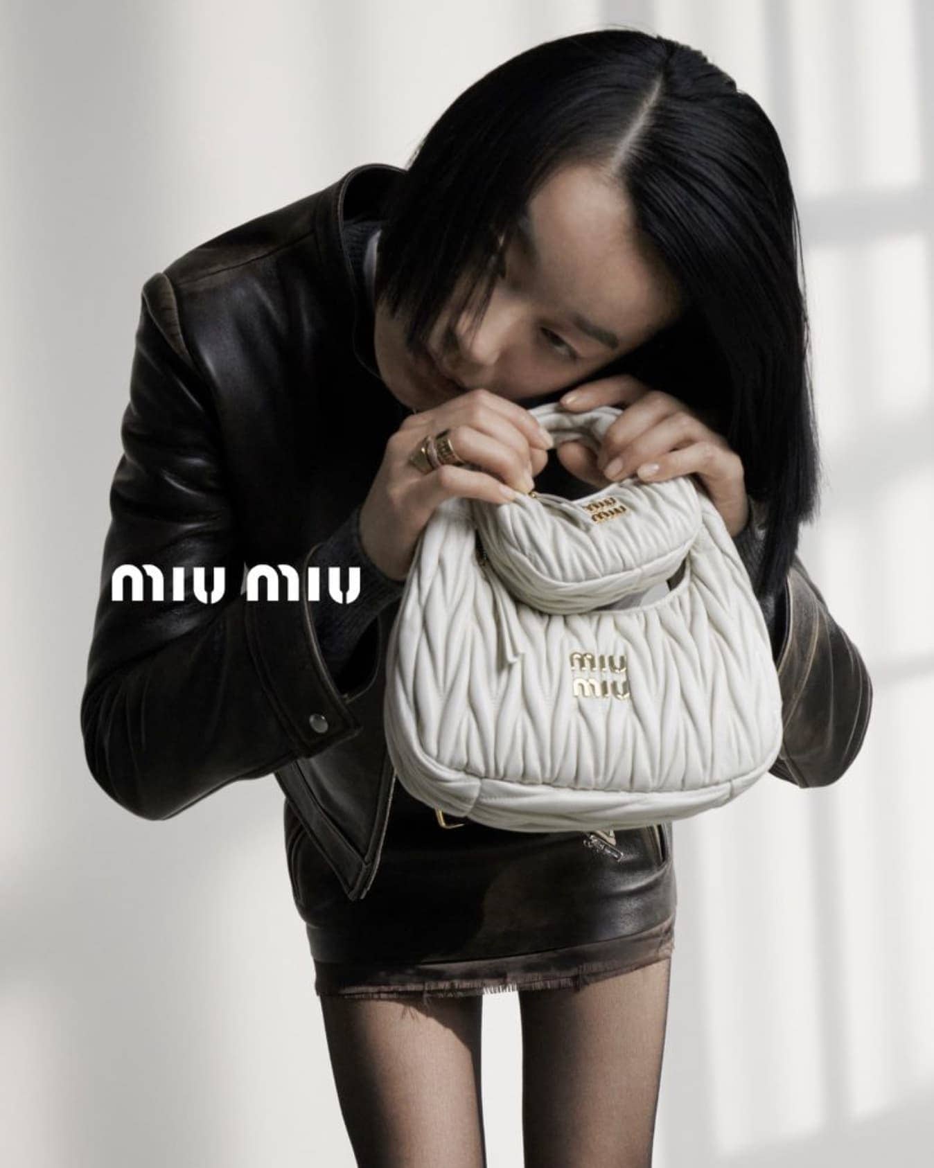 An adult female with short black hair poses indoors with a white leather purse in front of a grey backdrop, wearing a black leather jacket and skirt.