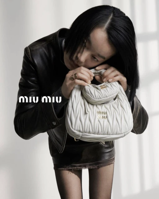An adult female with short black hair poses indoors with a white leather purse in front of a grey backdrop, wearing a black leather jacket and skirt.