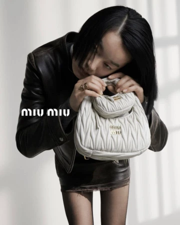 An adult female with short black hair poses indoors with a white leather purse in front of a grey backdrop, wearing a black leather jacket and skirt.