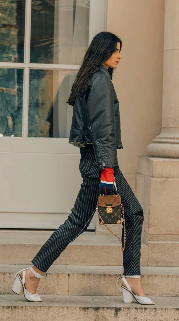 Fashionable woman walks on steps outdoors with a patterned Louis Vuitton handbag, wearing a black jacket and striped pants.