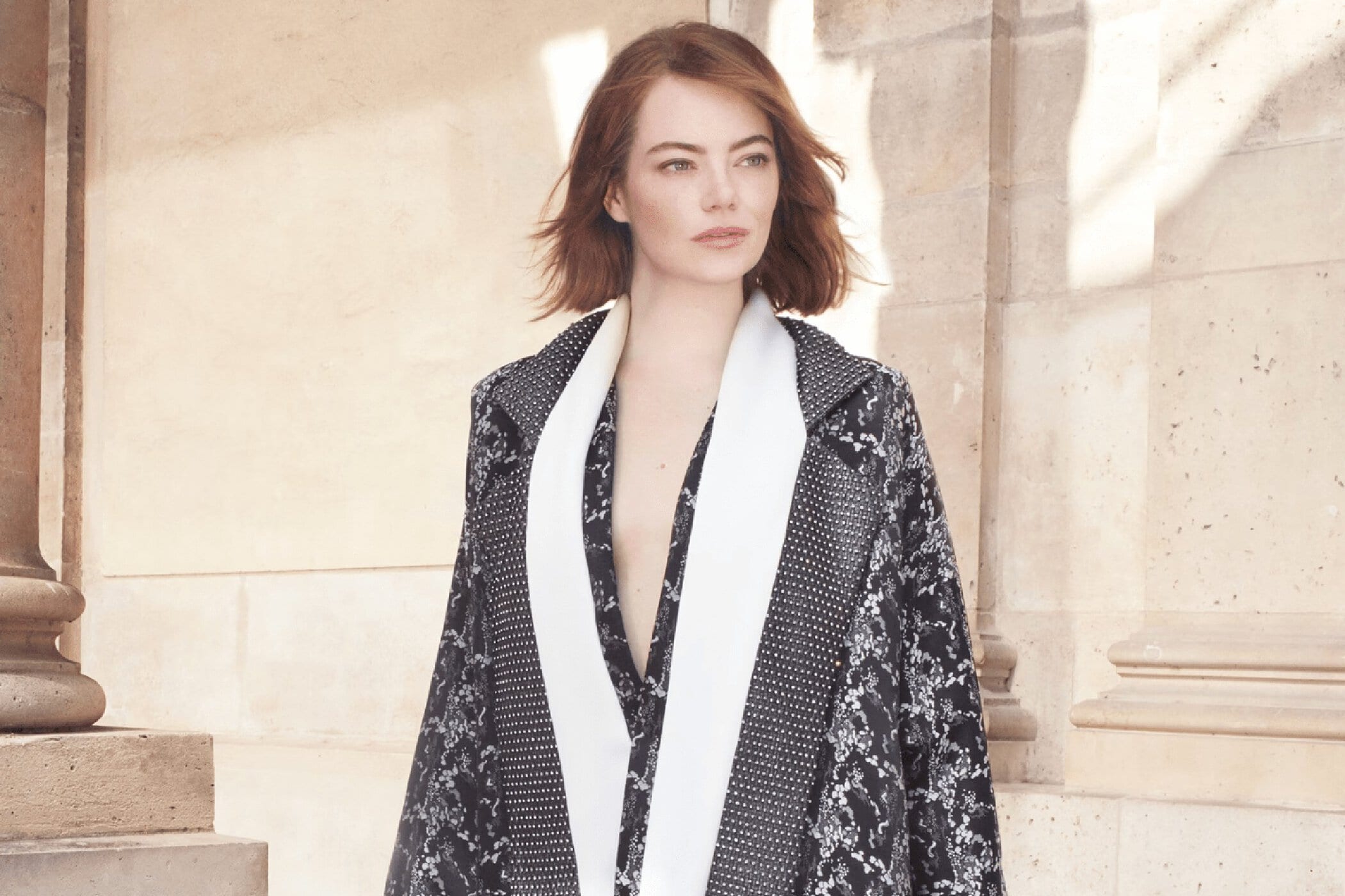 Adult woman with medium auburn hair wearing patterned coat with a white scarf poses in front of an off-white building in a portrait.