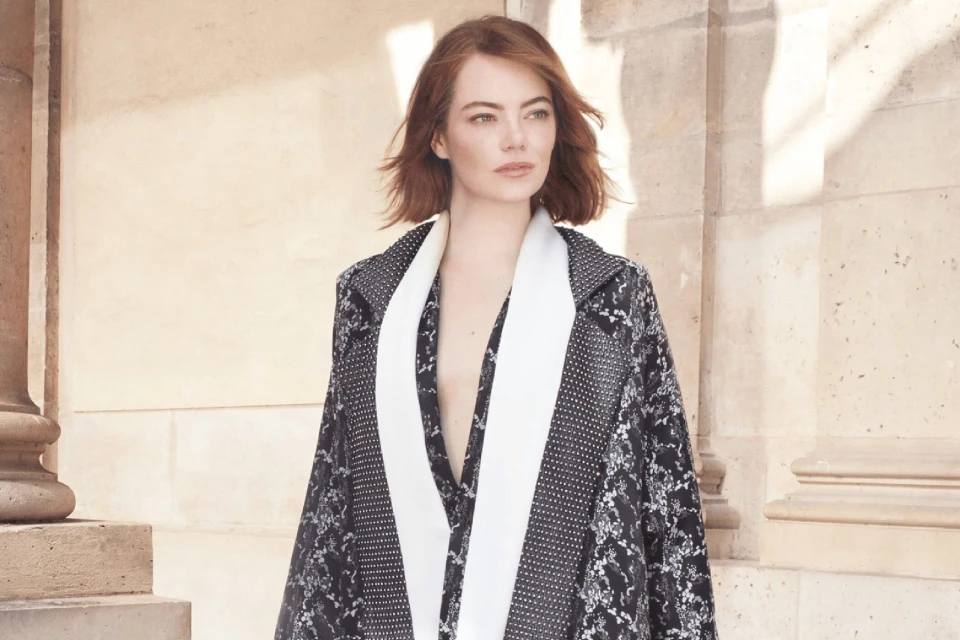 Adult woman with medium auburn hair wearing patterned coat with a white scarf poses in front of an off-white building in a portrait.