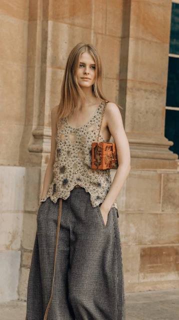 A slender, light-skinned woman with long blonde hair models a beaded top, wide leg trousers, and structured orange purse near a stone wall.