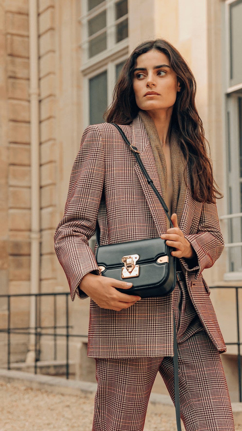 Adult female model poses in a suit, holding a purse, with a neutral expression in front of a building with architectural details.