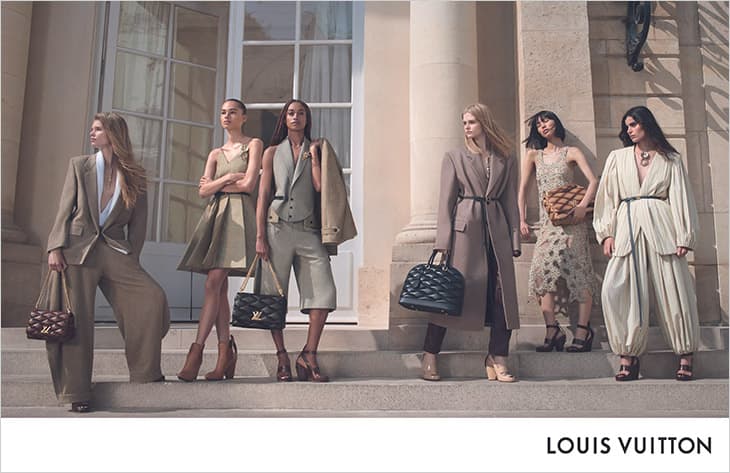 Group of six fashion models in neutral-toned outfits and bags standing on stairs outdoors for a Louis Vuitton fashion ad campaign.