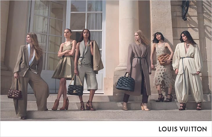 Group of six fashion models in neutral-toned outfits and bags standing on stairs outdoors for a Louis Vuitton fashion ad campaign.