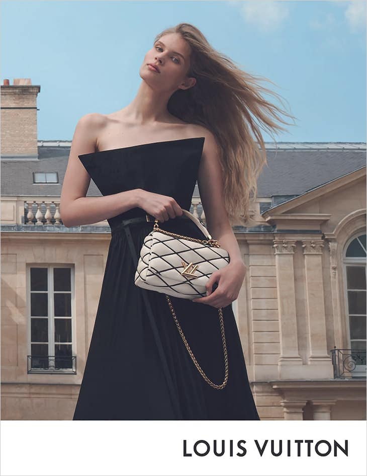 Fashion shot of a blond woman wearing a black dress and holding a white Louis Vuitton handbag in front of a cityscape backdrop.