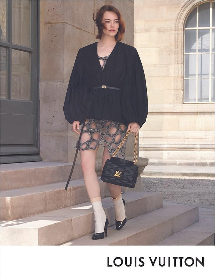 Emma Stone poses on stone steps with a black Louis Vuitton purse, wearing a black top, patterned skirt, and white socks with black heels.