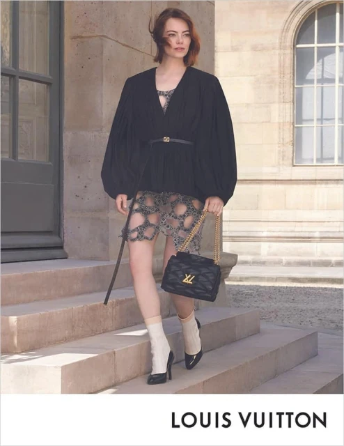 Emma Stone poses on stone steps with a black Louis Vuitton purse, wearing a black top, patterned skirt, and white socks with black heels.