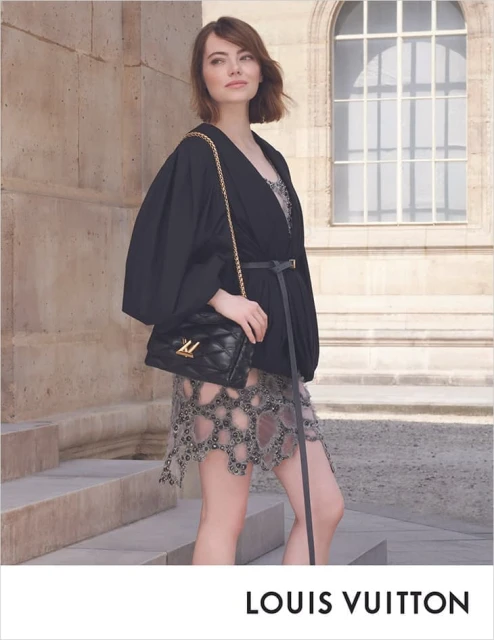 Emma Stone poses in front of a building wearing black jacket, jeweled dress, and carrying a quilted Louis Vuitton handbag with a gold chain.