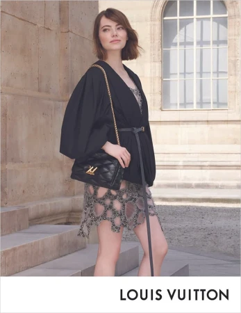Emma Stone poses in front of a building wearing black jacket, jeweled dress, and carrying a quilted Louis Vuitton handbag with a gold chain.