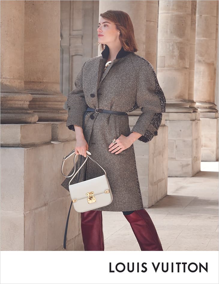 Fashion shot of a woman in a grey coat, maroon pants and a white bag, standing in front of stone columns and wearing luxury apparel.