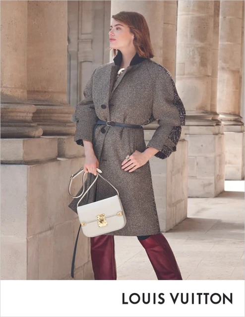 Fashion shot of a woman in a grey coat, maroon pants and a white bag, standing in front of stone columns and wearing luxury apparel.