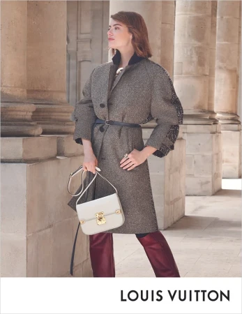 Fashion shot of a woman in a grey coat, maroon pants and a white bag, standing in front of stone columns and wearing luxury apparel.