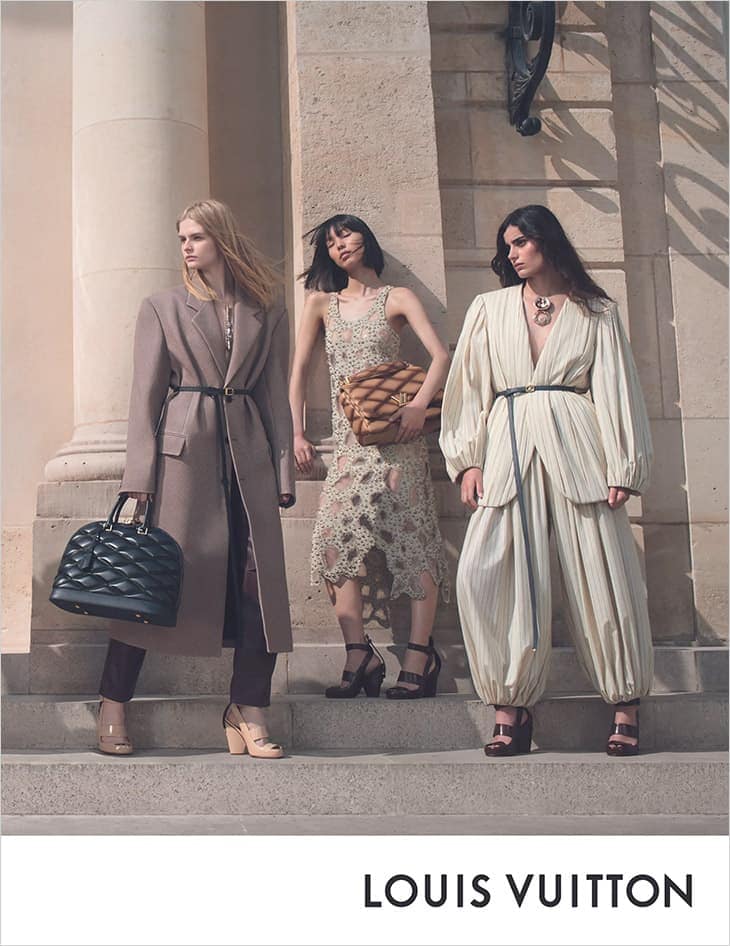 Three fashion models pose on stone steps in stylish outfits, featuring a coat, a dress, and a draped jumpsuit. Louis Vuitton logo visible.