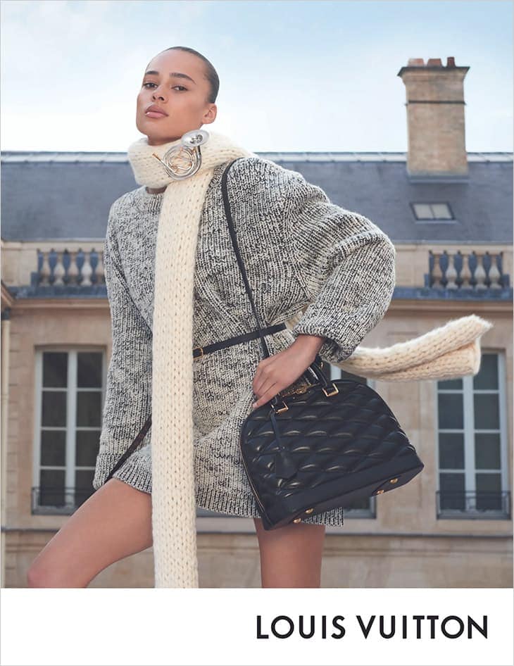 Fashion shoot featuring a woman modeling a black quilted handbag, wearing a grey patterned sweater dress and ivory scarf on a rooftop.