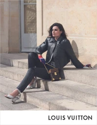 Model seated on outdoor stone steps in a black cropped leather jacket, dark textured trousers, white pumps, and a Louis Vuitton monogram bag.