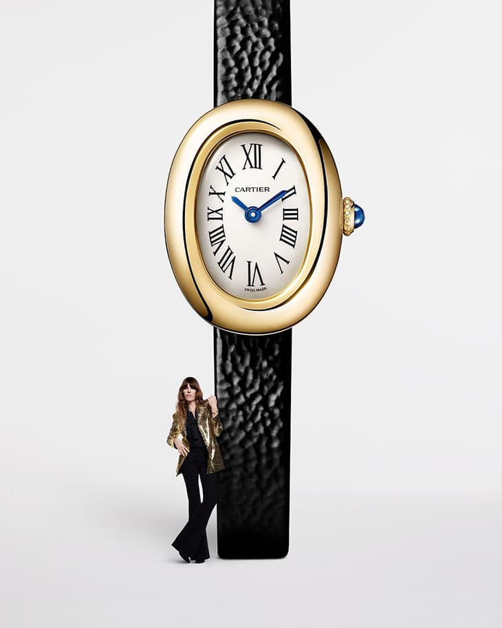 Wristwatch with a gold case, black strap, and white face displayed with a woman in a shiny blazer and black pants in studio lighting.