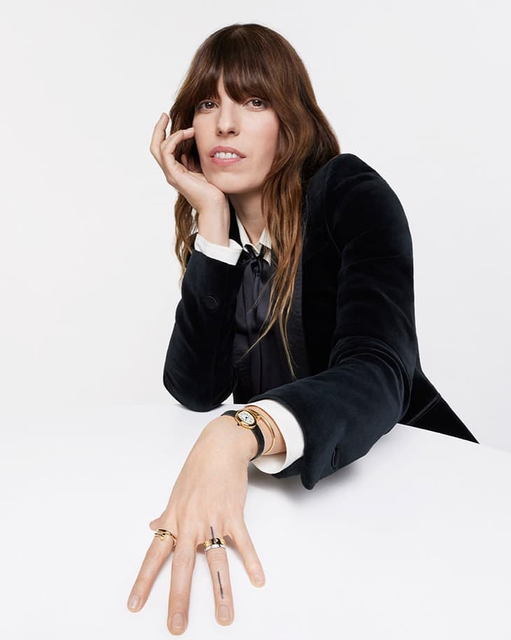 Close-up shot of a woman with long brown hair wearing a dark velvet blazer, jewelry, and a watch, posing against a white background.
