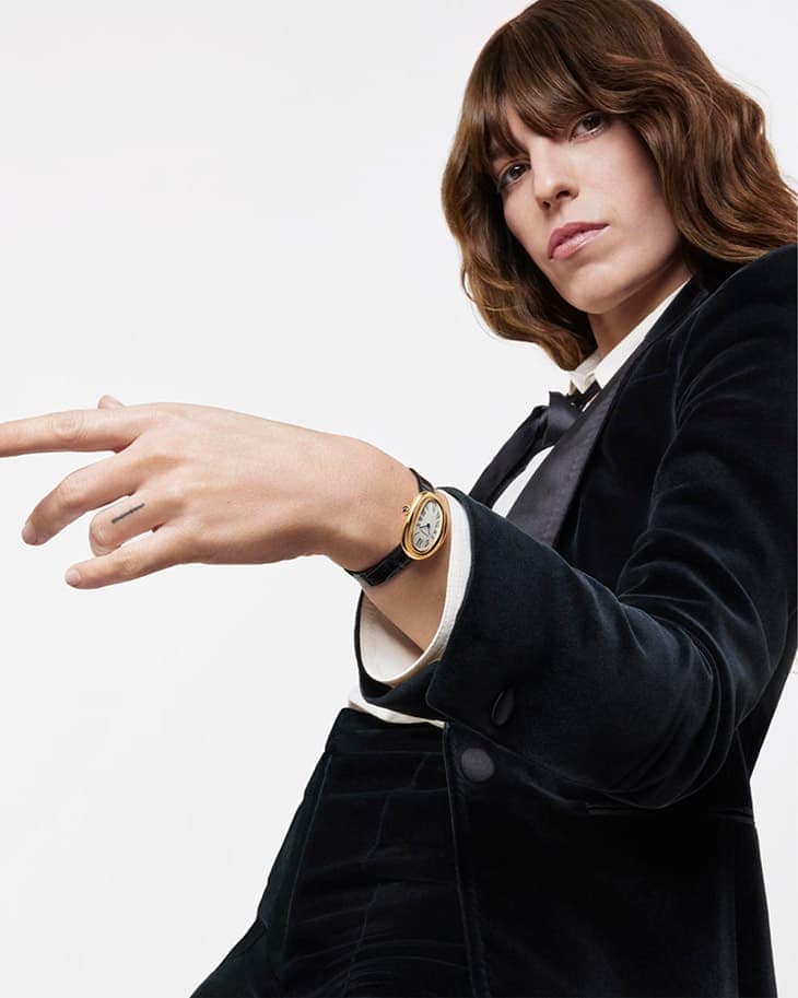 A woman in a black velvet suit and bow tie poses against a white backdrop wearing a gold watch with a black strap on her left wrist.