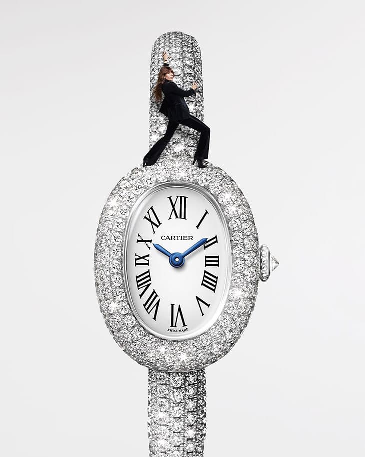 Close-up of a Cartier watch adorned with diamonds, featuring a woman in a black suit balancing on top, set against a white backdrop.