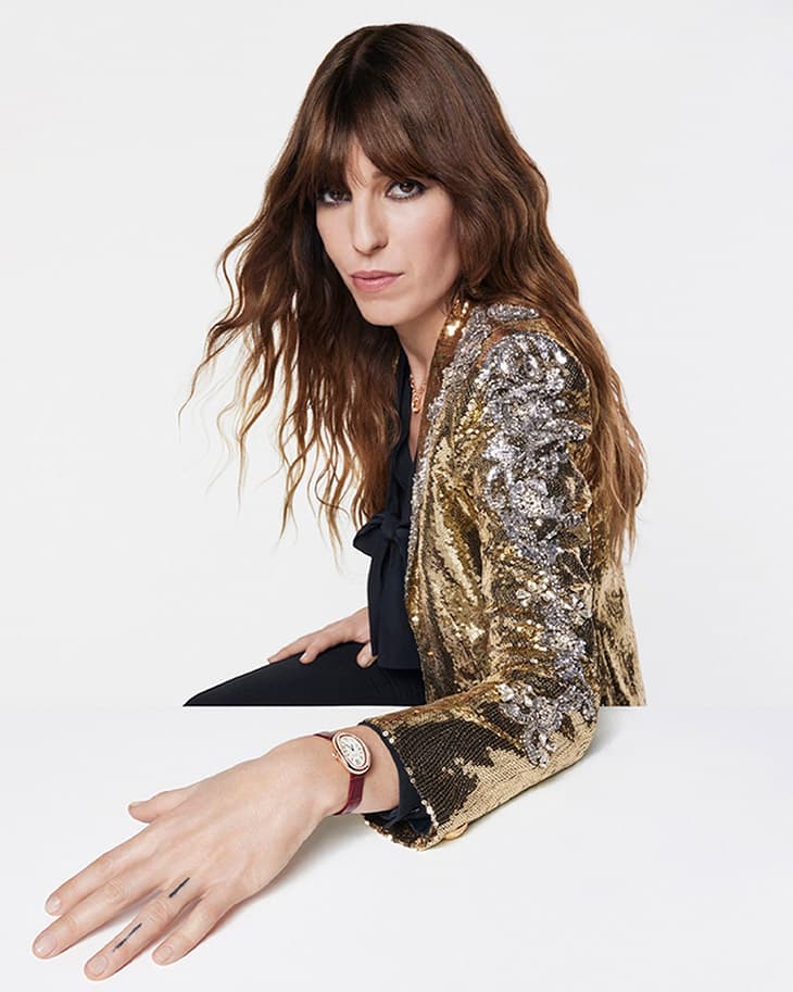 A brunette woman poses wearing a sequined gold jacket and a watch, set against a bright white background in a fashion-style studio portrait.