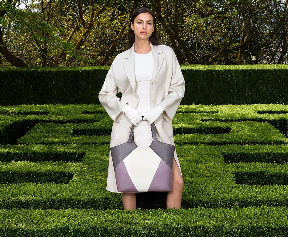 Fashion model Irina Shayk stands in a green hedge maze wearing an off-white coat and carrying a color block leather handbag.