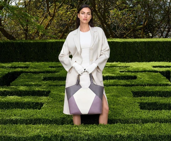 Fashion model Irina Shayk stands in a green hedge maze wearing an off-white coat and carrying a color block leather handbag.
