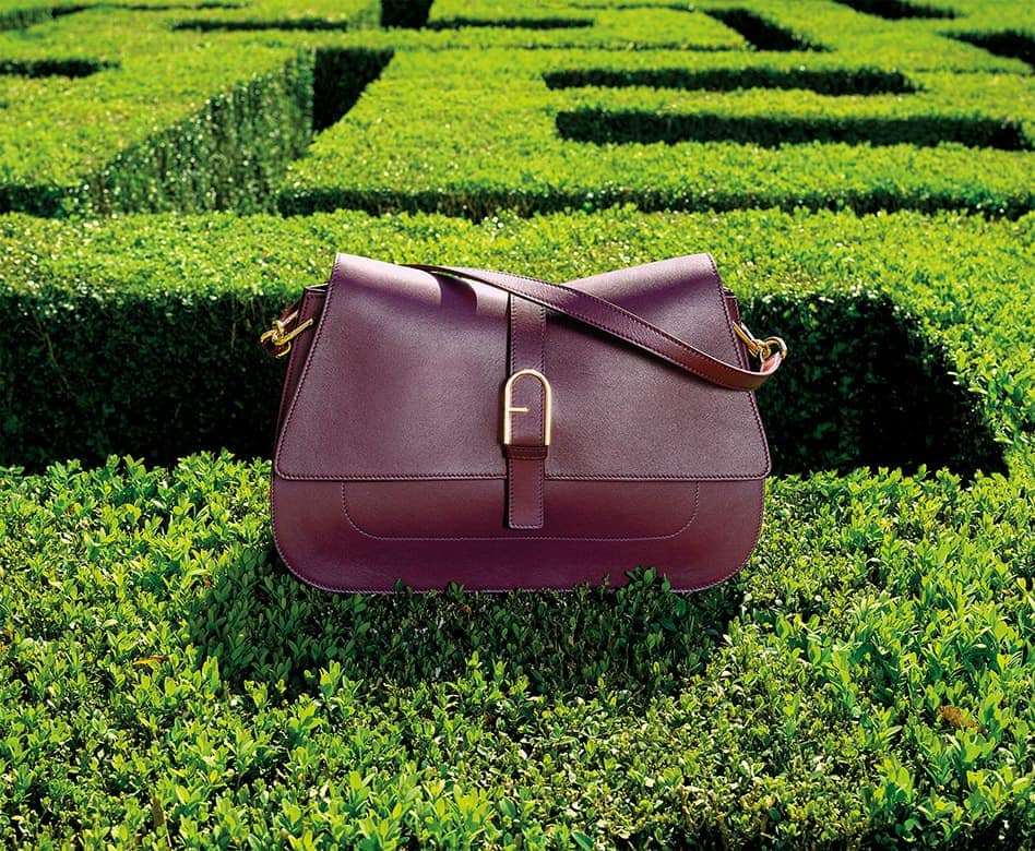 A brown leather shoulder bag with gold hardware sits on a trimmed hedge against the backdrop of a neatly manicured hedge maze.