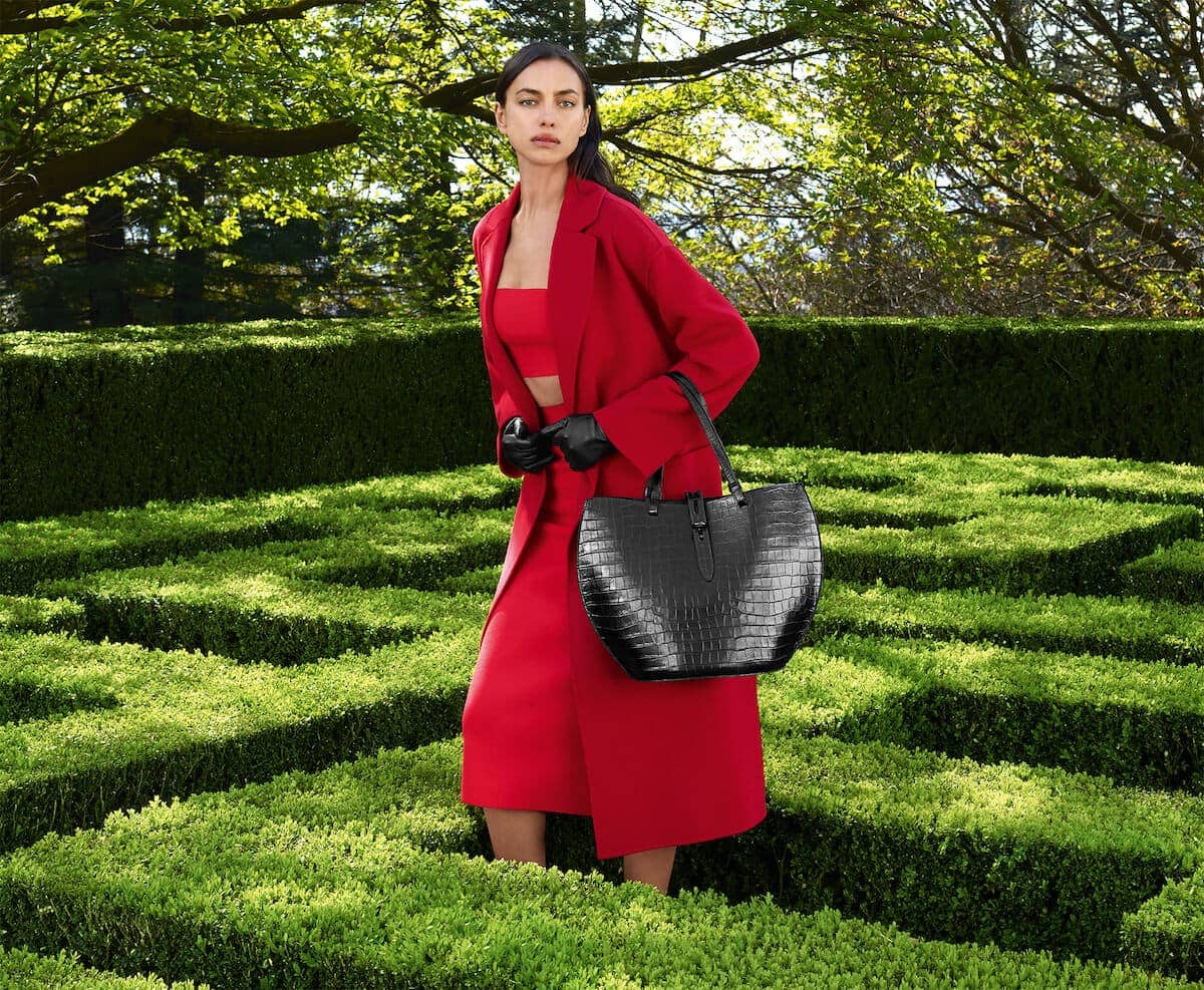 Elegant woman in a red outfit and a black leather handbag standing in a green topiary maze on a sunny day.