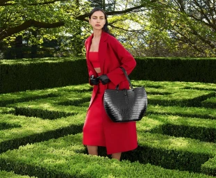 Model Irina Shayk wearing a red coat and skirt holding a black handbag in a garden maze for Furla Fall 2023 ad campaign.