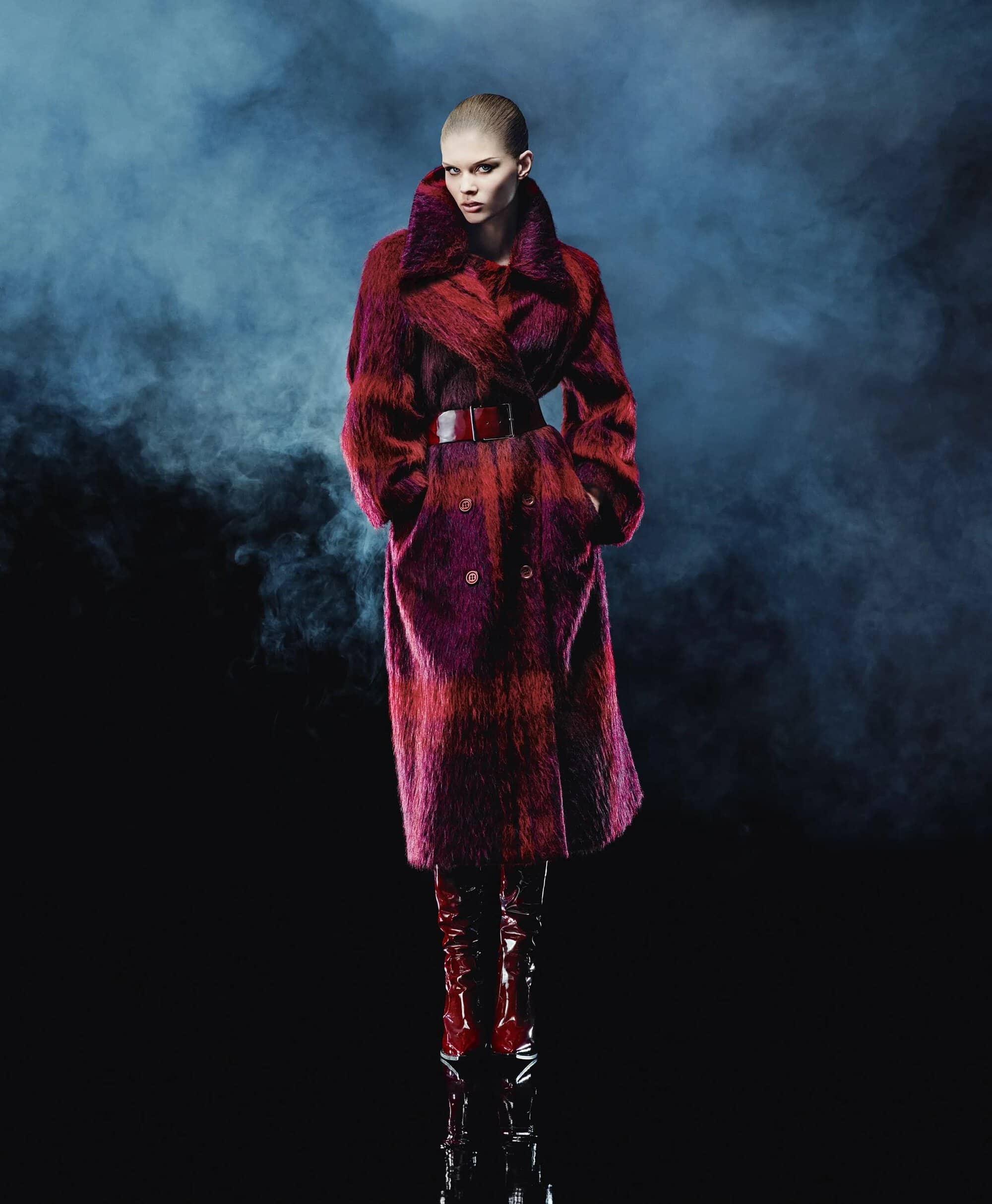 Fashion model posing in a red plaid coat, belt, and high boots against a dark smoky backdrop in a studio fashion shot.