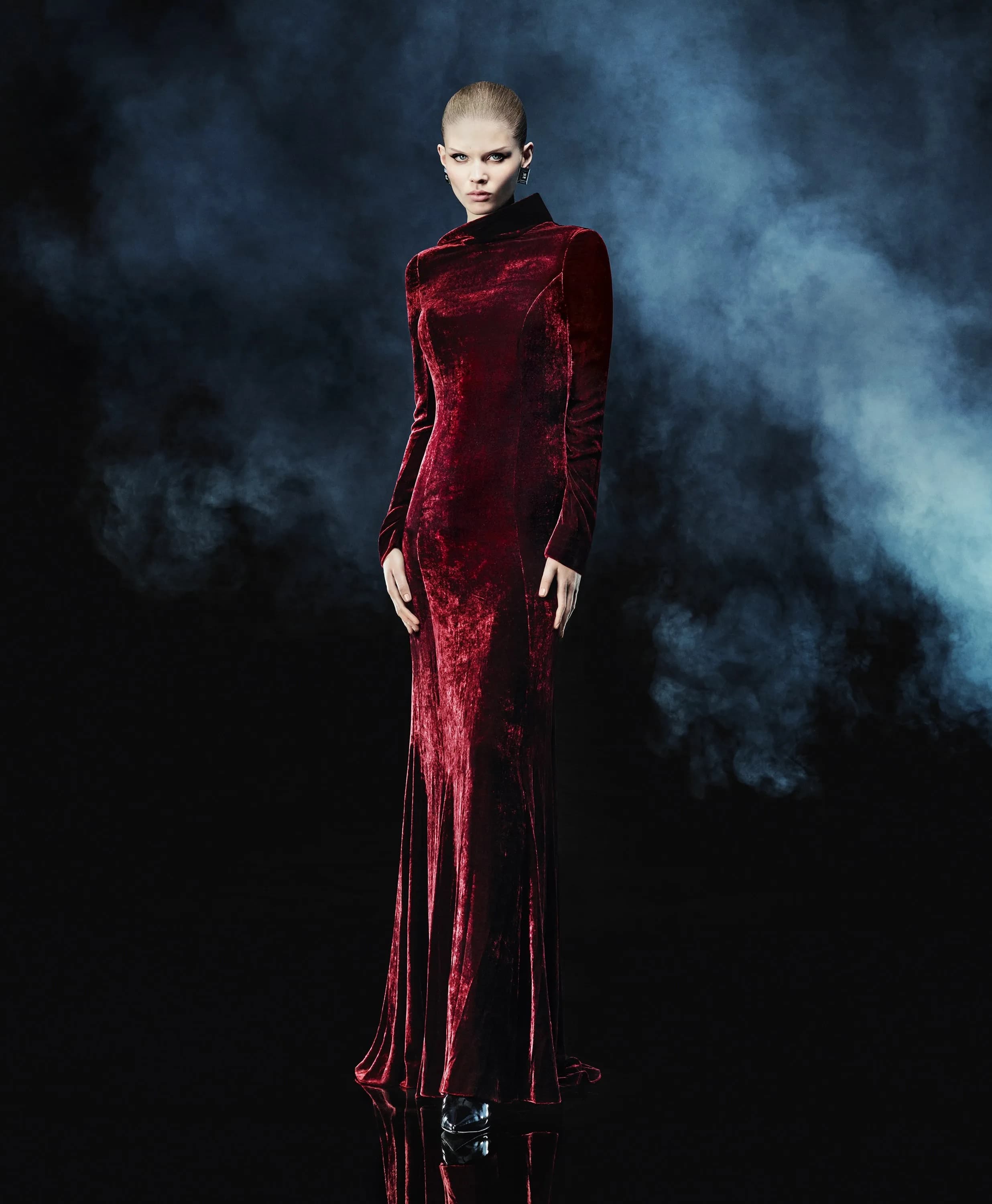 A woman stands in a red velvet gown against a smoky black background, showcasing fashion and formal wear.
