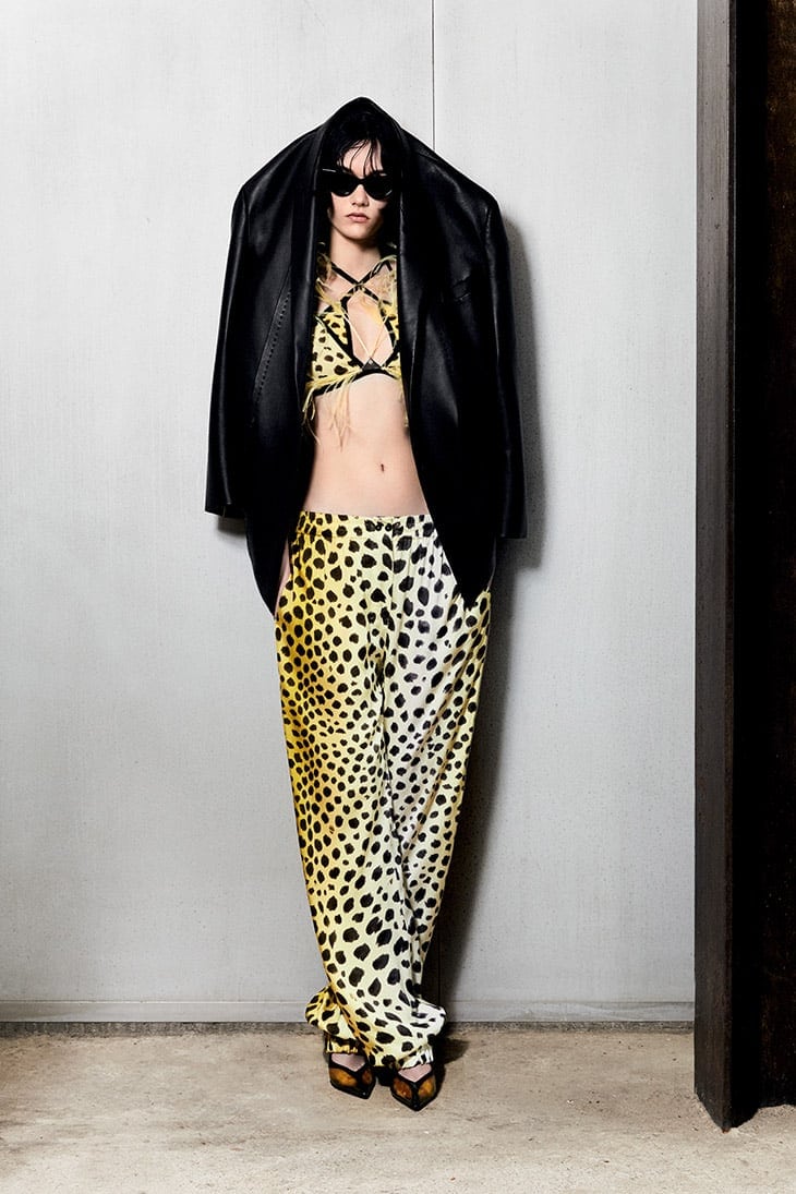 Fashion model poses in a cheetah print top and pants with a black leather jacket against a gray wall in a full-length shot.