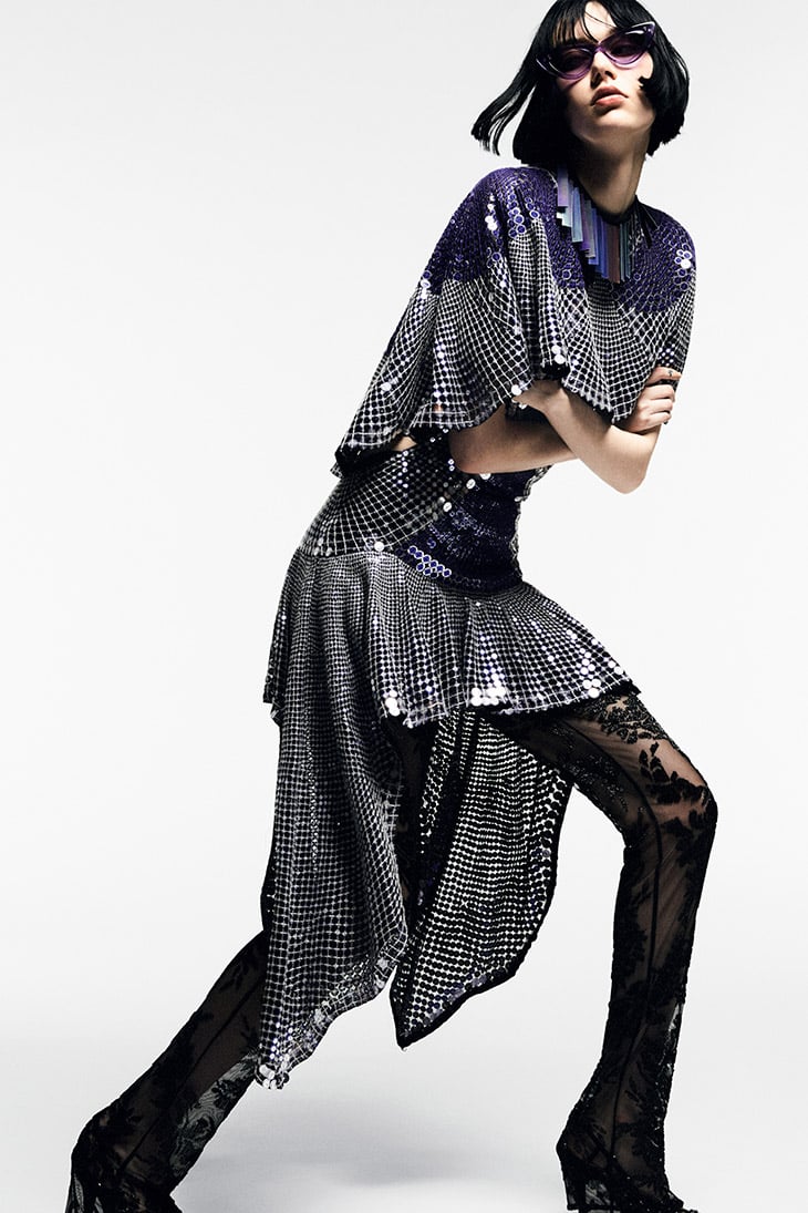 Fashionable woman strikes a pose wearing a silver and blue sequined dress, lace tights, and purple sunglasses in a studio shot.