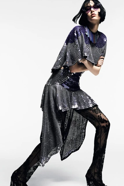 Fashionable woman strikes a pose wearing a silver and blue sequined dress, lace tights, and purple sunglasses in a studio shot.