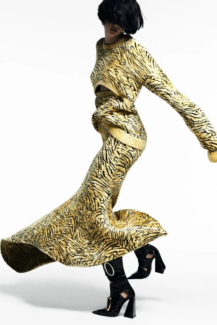 Woman in a patterned skirt and sweater with black heeled boots, striking a dynamic pose against a white background in a fashion editorial.