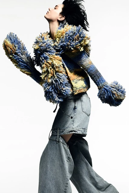 A woman in a blue and yellow yarn jacket and light-wash denim is posed against a white background in a studio shot, full body.