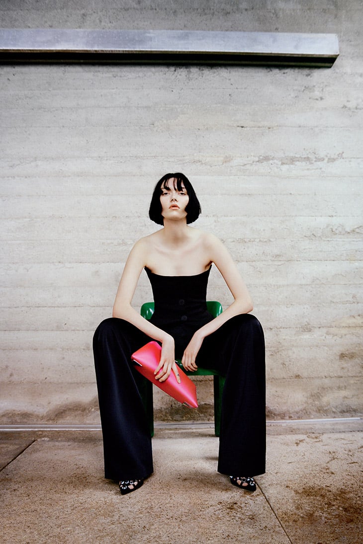 Model Steinberg seated on a green chair in a black strapless corset and wide-leg trousers, holding a hot pink clutch.