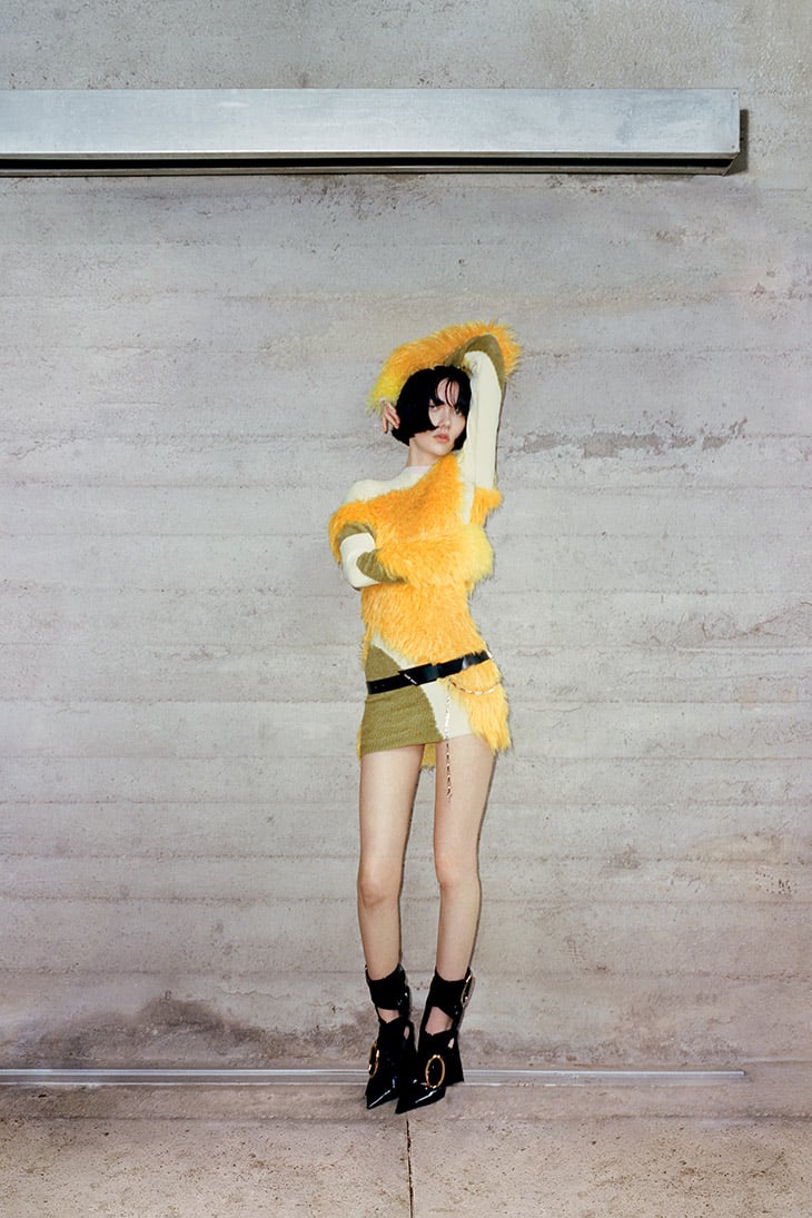 A person in a unique yellow and green outfit poses in front of a grey wall with a metal bar overhead, showcasing fashion and style.