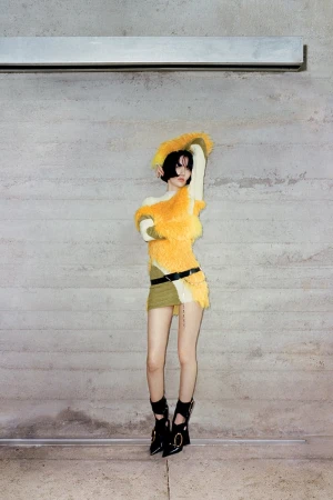 A person in a unique yellow and green outfit poses in front of a grey wall with a metal bar overhead, showcasing fashion and style.