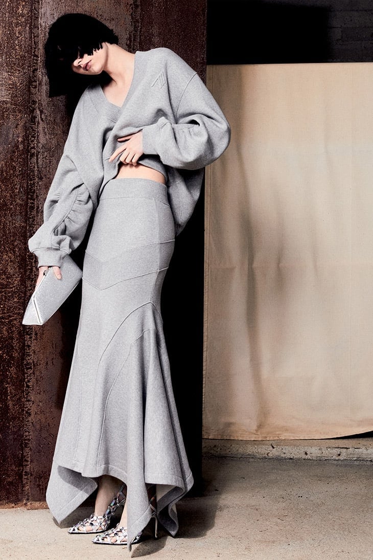 An adult woman poses against a textured wall wearing a grey, oversized sweater and a form-fitting, long grey skirt, also holding a clutch purse.