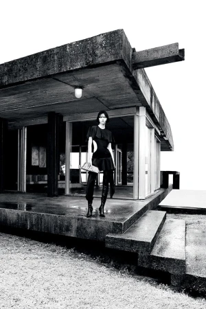 Model Steinberg poses in a black cutout dress and high boots on the concrete terrace of a brutalist building in black and white.