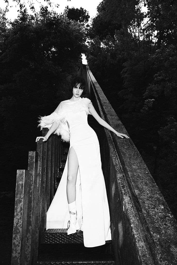 A black and white fashion portrait of a model in a long white dress with feathers on a wooden walkway surrounded by trees.