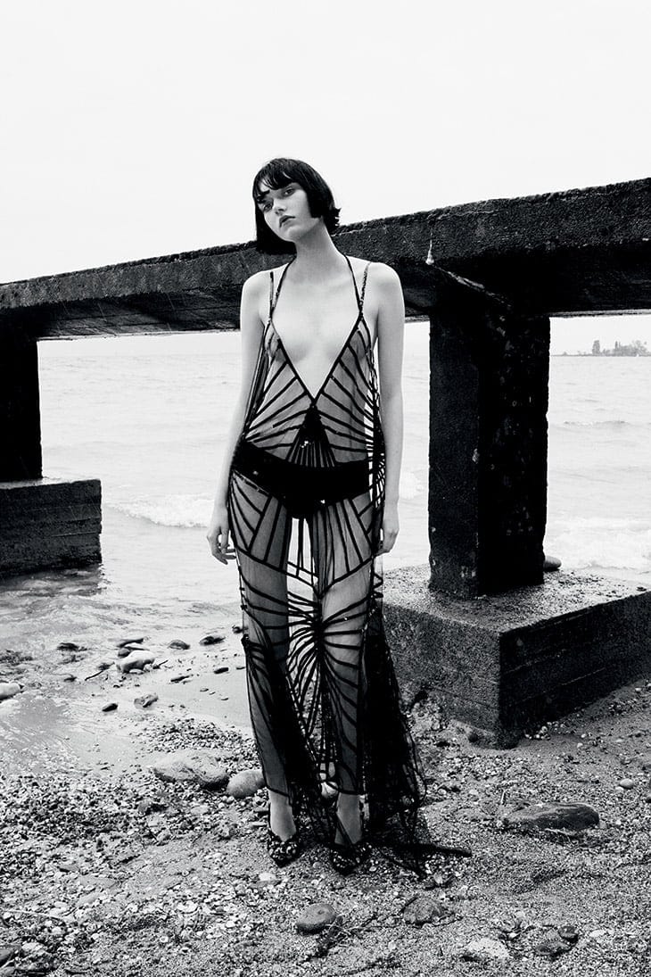 A model with a short bob haircut wears a black, sheer, geometrically patterned gown while standing on a rocky beach by a concrete pier.