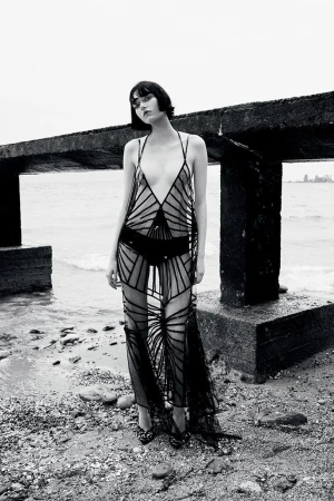 A model with a short bob haircut wears a black, sheer, geometrically patterned gown while standing on a rocky beach by a concrete pier.