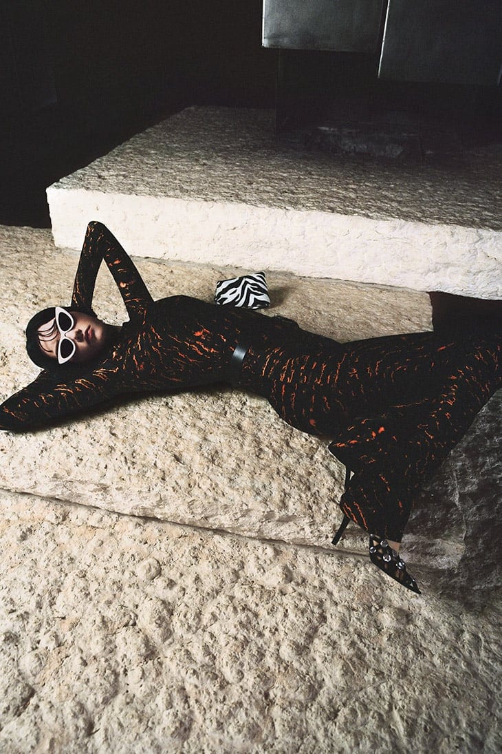 A female fashion model with sunglasses is lying on a stone surface wearing a black and orange printed dress in a studio.