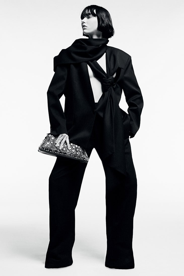 Fashion editorial photo of a woman posing in a black blazer and trousers, holding a beaded clutch in a white photo studio.