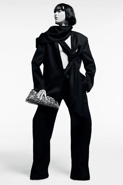 Fashion editorial photo of a woman posing in a black blazer and trousers, holding a beaded clutch in a white photo studio.